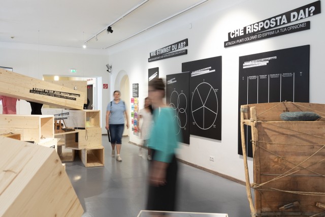 Man sieht eine Ausstellung im Volkskunstmuseum mit Menschen, die sich bewegen und deshalb verschwommen sind.